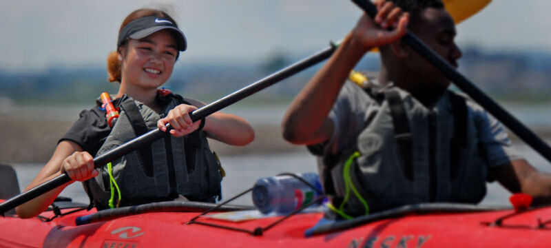The image shows two people kayaking on a sunny day. The person in the front is a young woman wearing a visor and a life vest, smiling at the camera. The other person is a young man, also wearing a life vest, paddling in the same direction. The kayak is red.